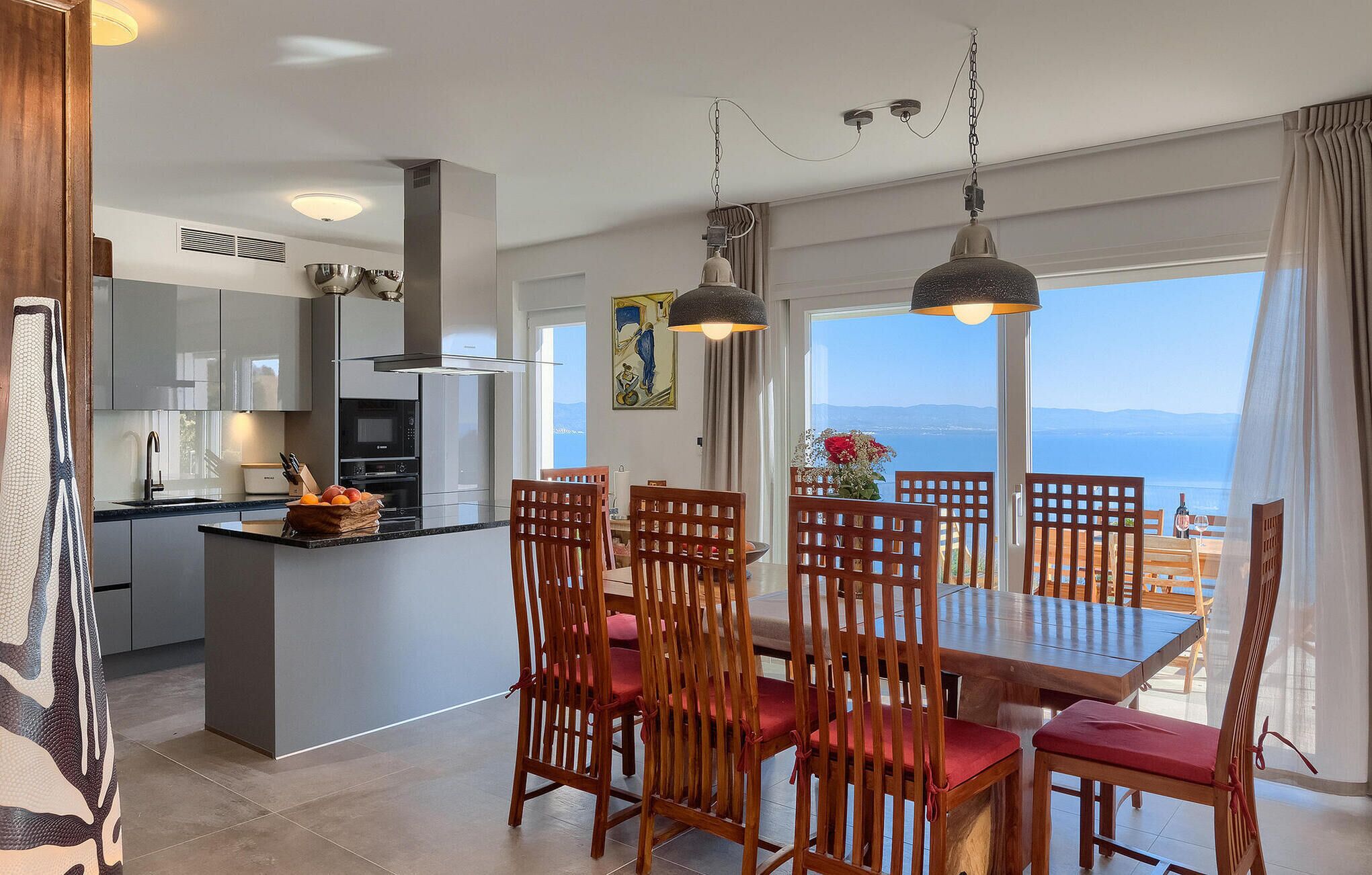 Una cocina moderna con una isla central, comedor con sillas y mesa de madera, y vista al mar a través de amplios ventanales.