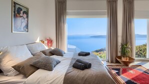 Ein Schlafzimmer mit einem großen Bett, beige farbenen Vorhängen, Meerblick und einem Bild an der Wand.