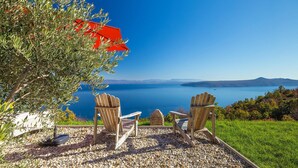 Eine Holzterrasse mit zwei Adirondack-Stühlen, die auf einen See ausgerichtet sind, einem roten Sonnenschirm und Blick auf ferne Berge.