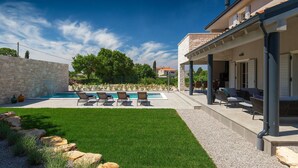 Ein Poolbereich mit Steinmauer, Rasenfläche und überdachter Terrasse mit Gartenmöbeln.
