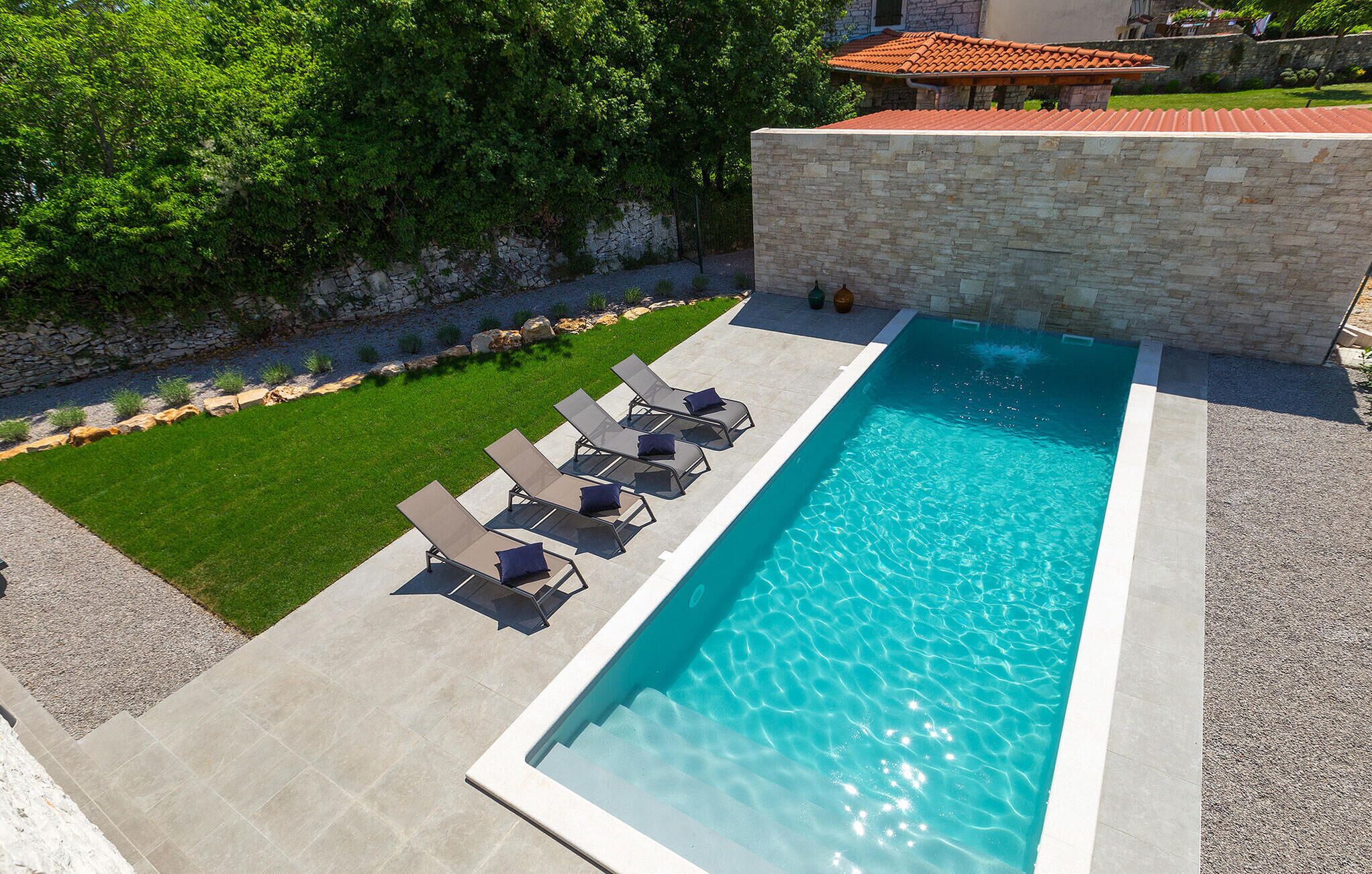 A rectangular swimming pool with clear blue water, surrounded by a stone wall and a grassy area with lounge chairs.