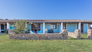 A two-story building with a stone facade, a covered patio with seating, and a clear blue sky. - La Casa Azzurra with shared Pool (Tertenia)