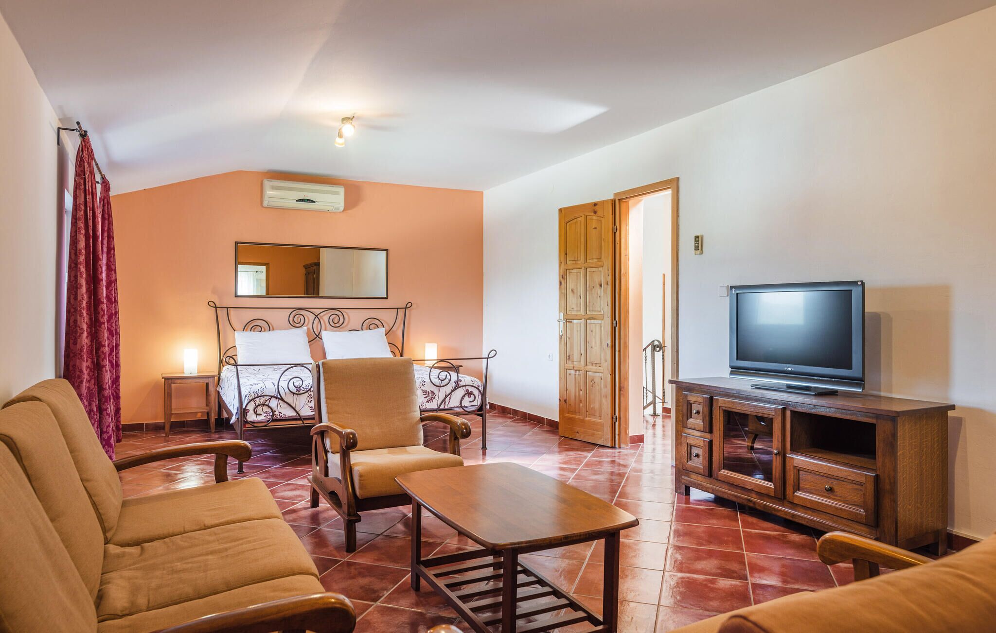 A living room with a sofa, armchairs, a coffee table, a TV on a wooden cabinet, a bed with a headboard, a mirror, and a ceiling fan.