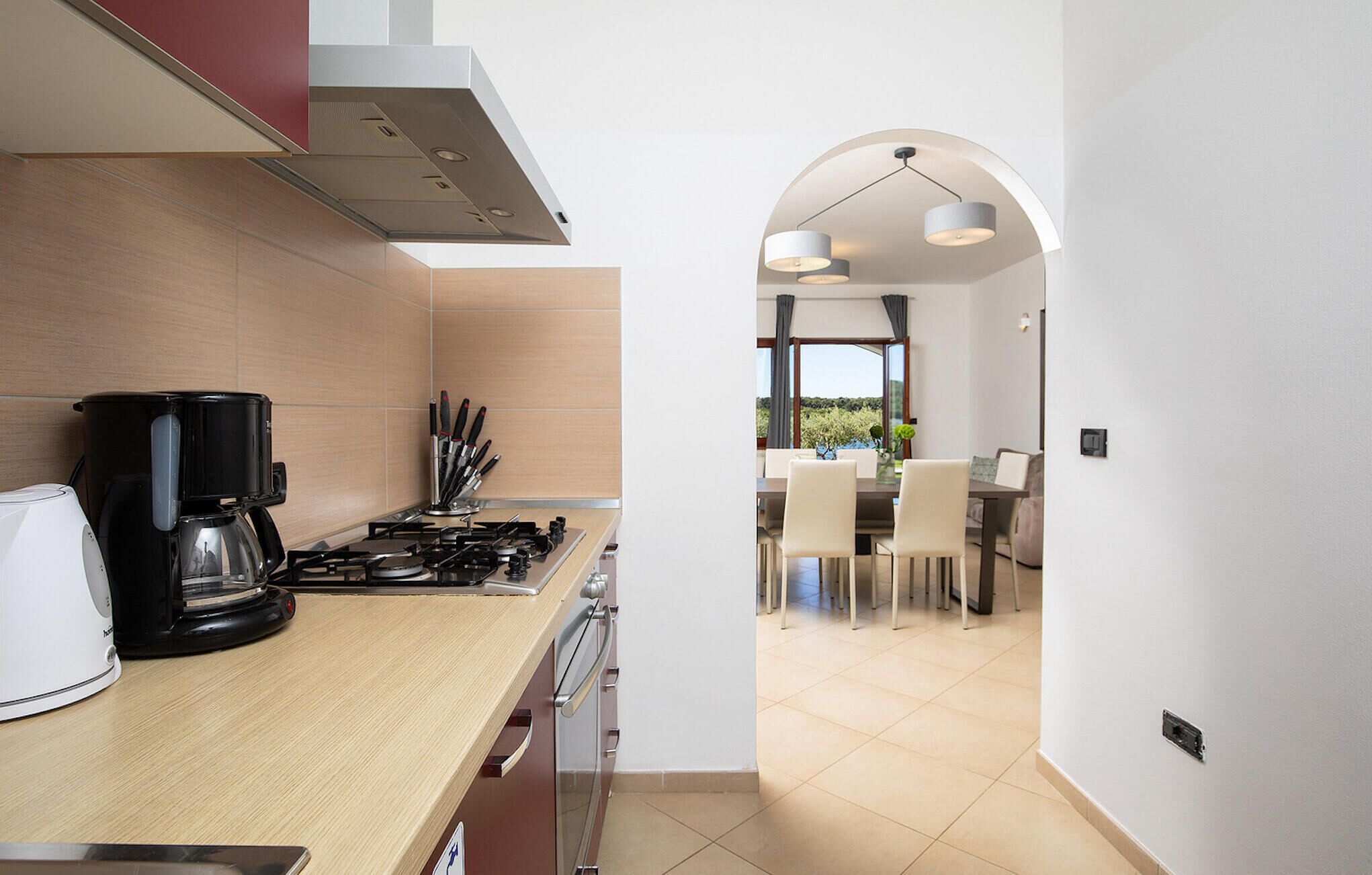A modern kitchen with a coffee maker, stove, and knives. There is a dining area with a table and chairs, and a view of the outdoors.