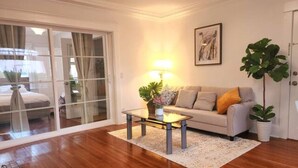 A living room with a glass door leading to a balcony, a white sofa with a yellow pillow, a glass coffee table with a plant, and a large green plant in a white pot.