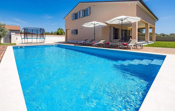A rectangular swimming pool with a clear blue water surface, surrounded by a wooden deck. There are lounge chairs and umbrellas on the deck, and a building with a beige facade and blue shutters in the background. - Stunning home in Pula (Pula)