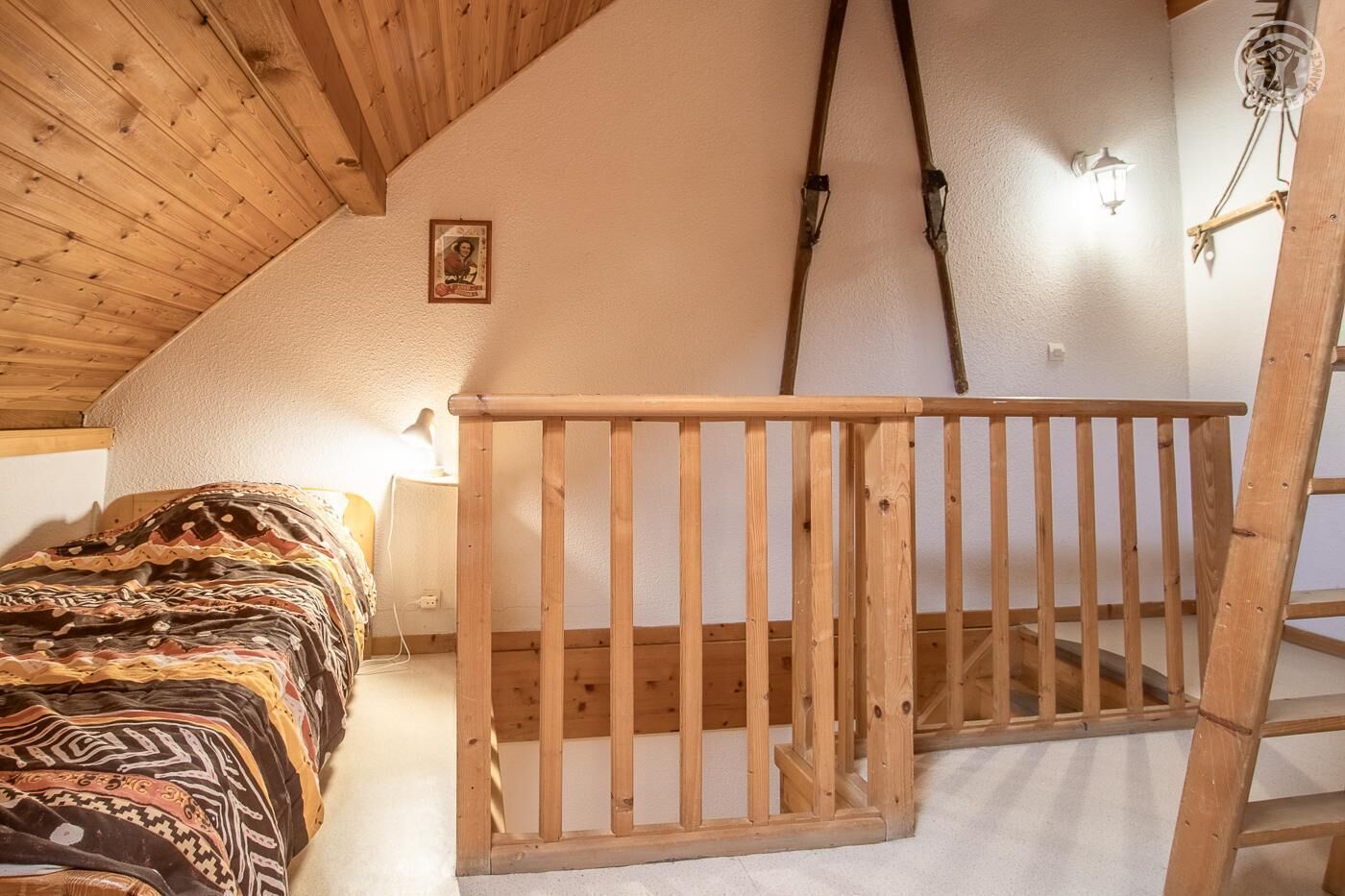 A bedroom with a wooden bunk bed, a ladder, a wall-mounted light fixture, and a framed picture on the wall.