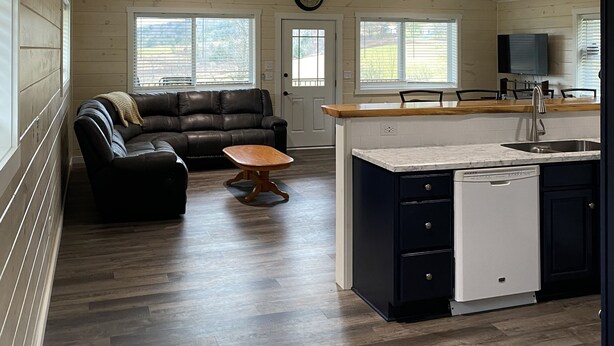 A spacious living area with a dark leather sofa, a wooden coffee table, and a kitchen with a white dishwasher and dark cabinets. - Family friendly cottage, covered deck, close to lake to fish, swim, and boat (Whitney Point)