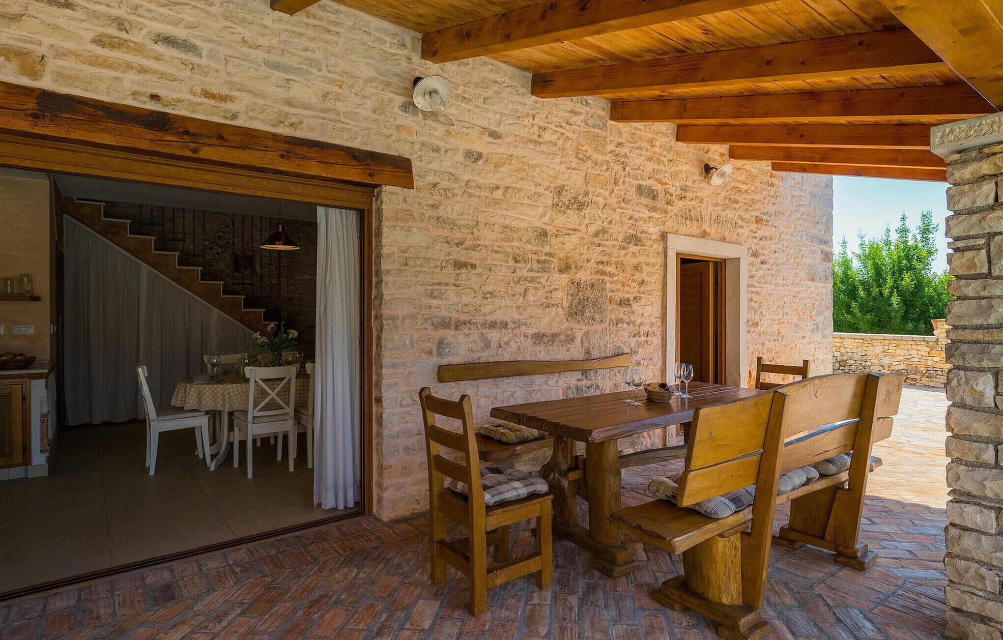 A rustic stone building with a wooden table and benches, a staircase, and a view of greenery outside.