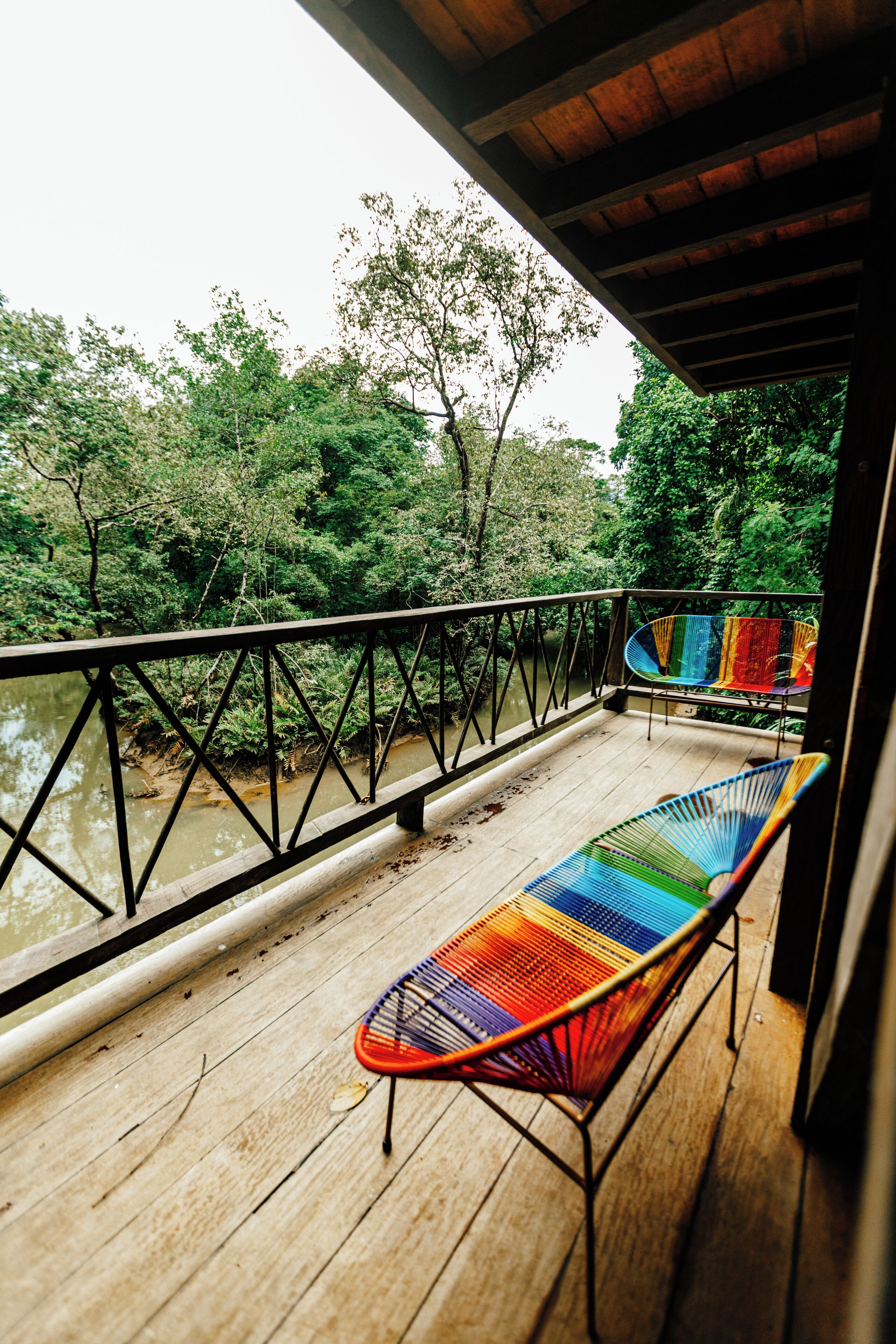 A wooden deck with a metal railing overlooking a river, with two colorful chairs and a hammock.
