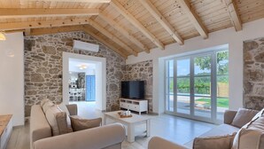 A spacious living area with stone walls, a wooden ceiling, a television on a white cabinet, and large windows offering a view of the outdoors.