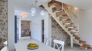 A modern kitchen with a white table, chairs, and a fruit bowl. A wooden staircase with a stone wall backdrop leads to another room.