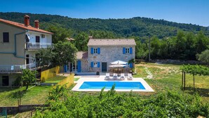 A two-story stone house with a swimming pool, surrounded by greenery and hills.