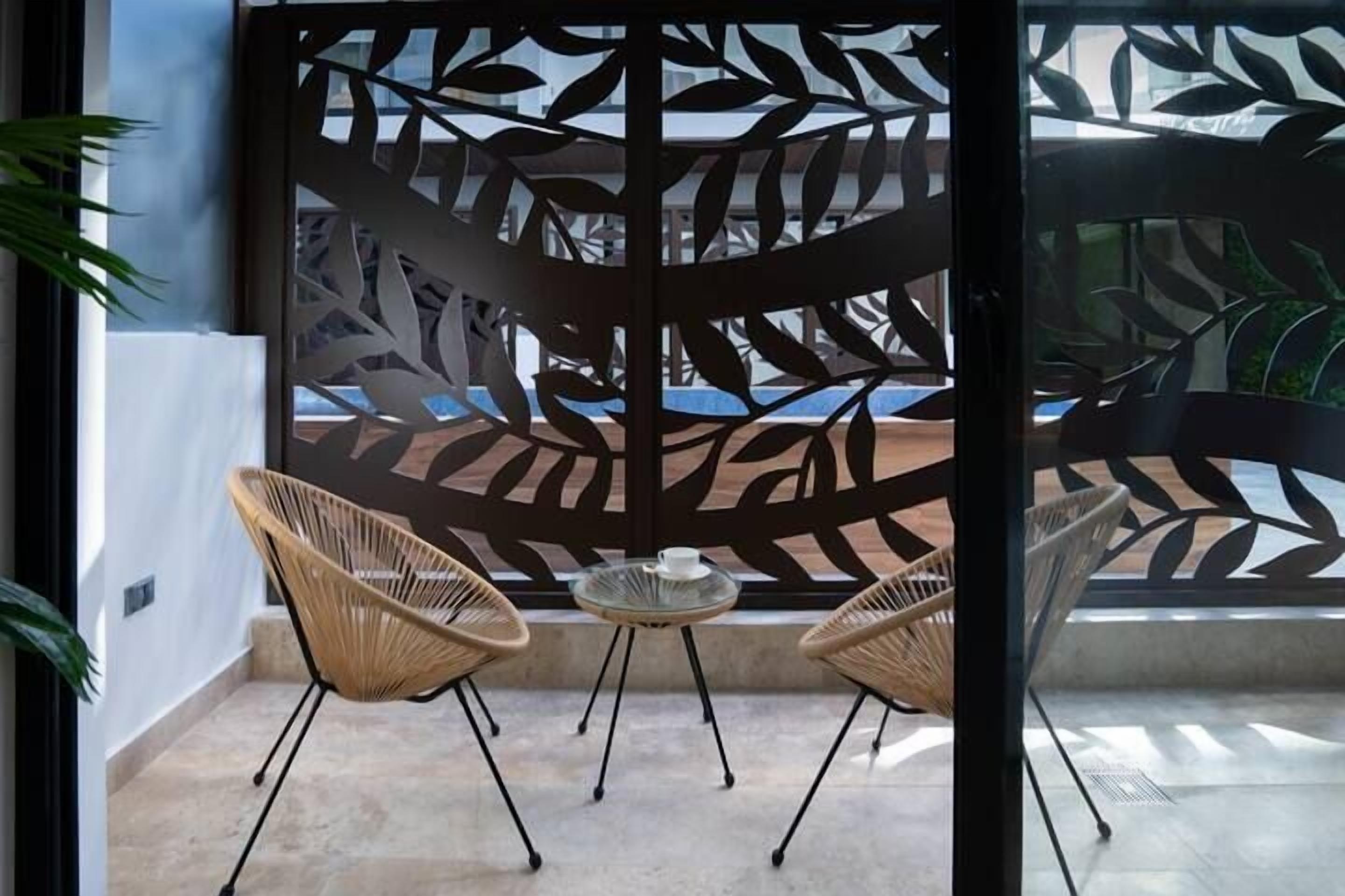 A balcony with two wicker chairs and a small glass table, a decorative metal partition with leaf patterns, and a view of a building in the background.