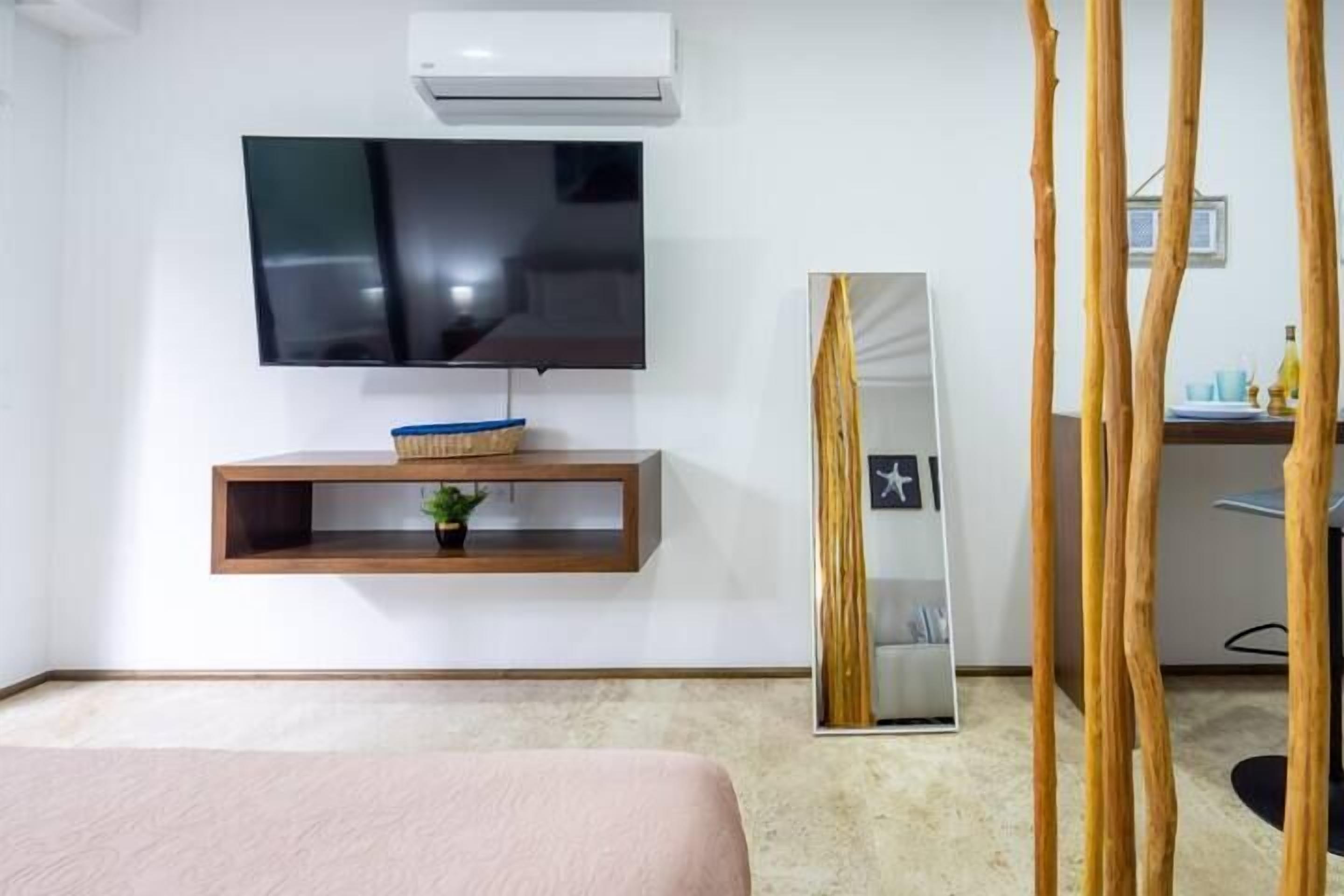 A modern hotel room with a flat-screen TV mounted on the wall, a wooden shelf with a plant, a large mirror, and a bed with a pink bedspread.