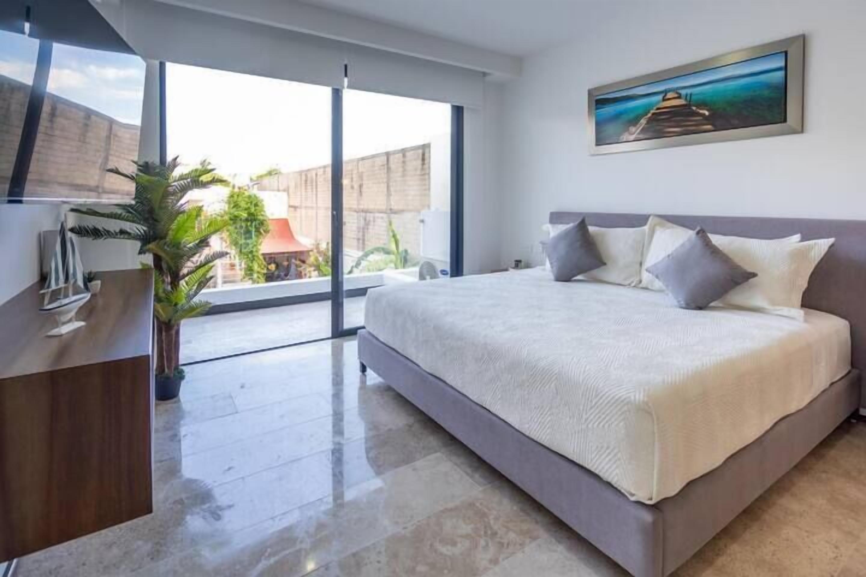 A modern bedroom with a large bed, a desk, and a sliding glass door leading to an outdoor area.