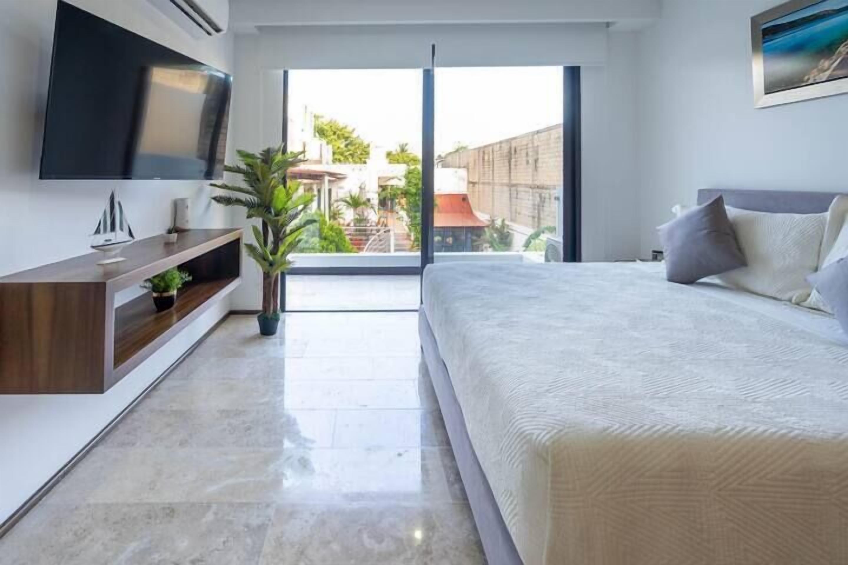 A modern bedroom with a large bed, a TV mounted on the wall, a small plant, and a view of the outside.