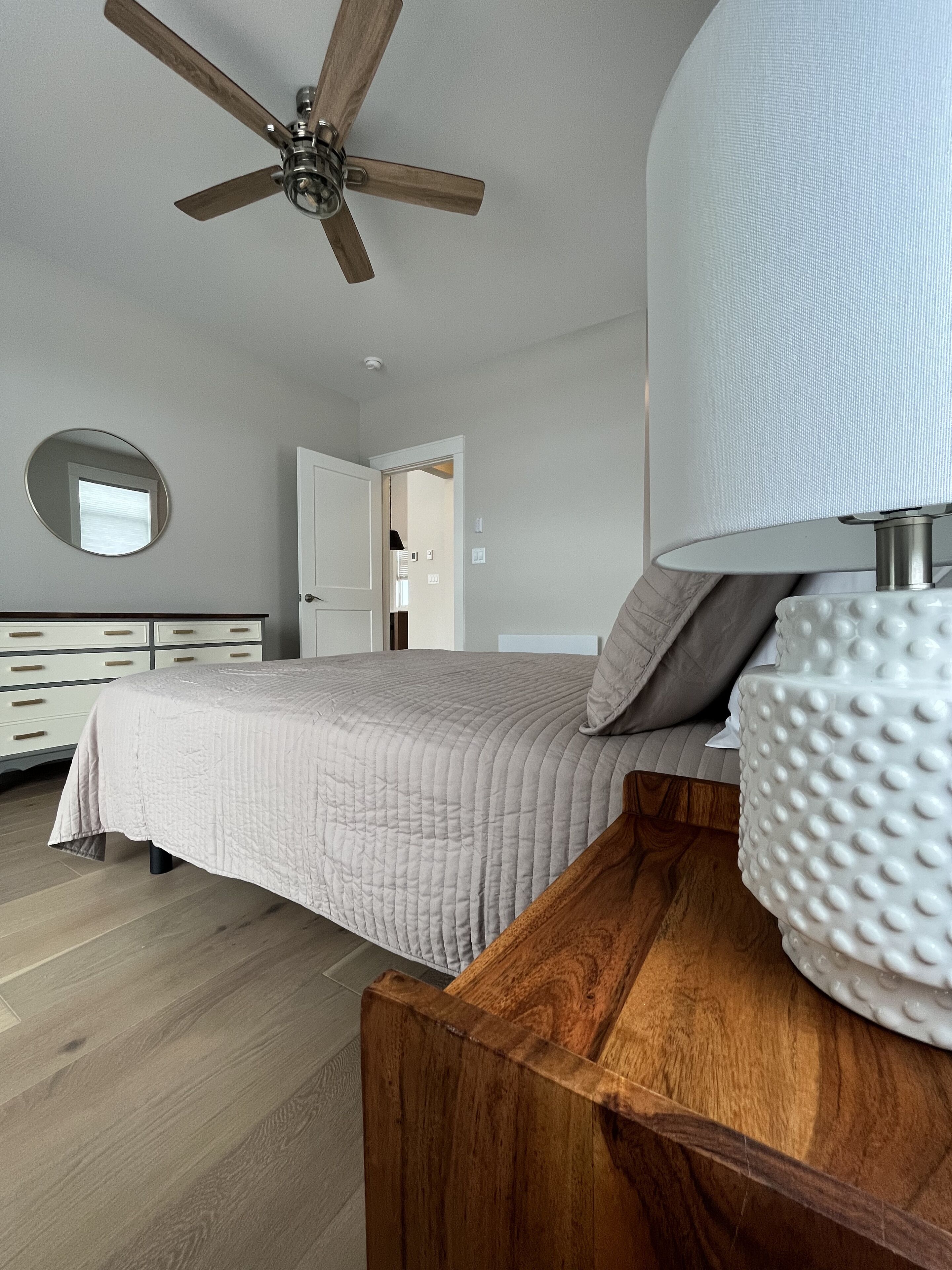 A bedroom with a bed, a wooden nightstand, a ceiling fan, a round mirror, and a door leading to another room.