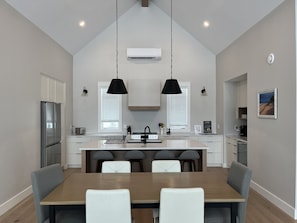 A modern kitchen with a central island, pendant lights, and a dining area with a wooden table and chairs. - Modern PEI Cottage with Stunning River Views - Near Cavendish (Clinton)
