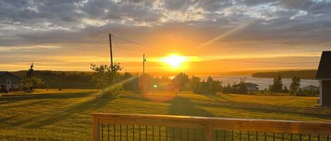 Une terrasse en bois avec une rambarde, donnant sur un champ verdoyant et une étendue d’eau au coucher du soleil.