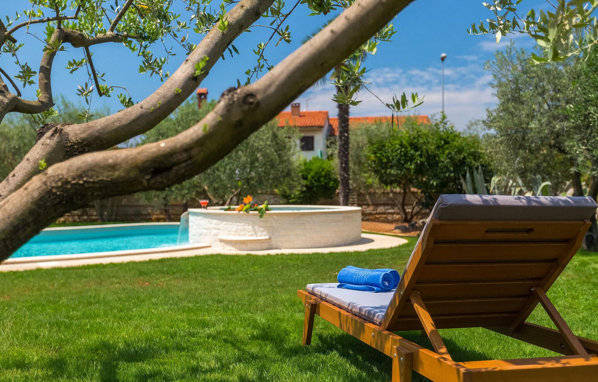 A wooden lounge chair with a cushion and a towel, a swimming pool with a stone edge, and a stone fountain with flowers.