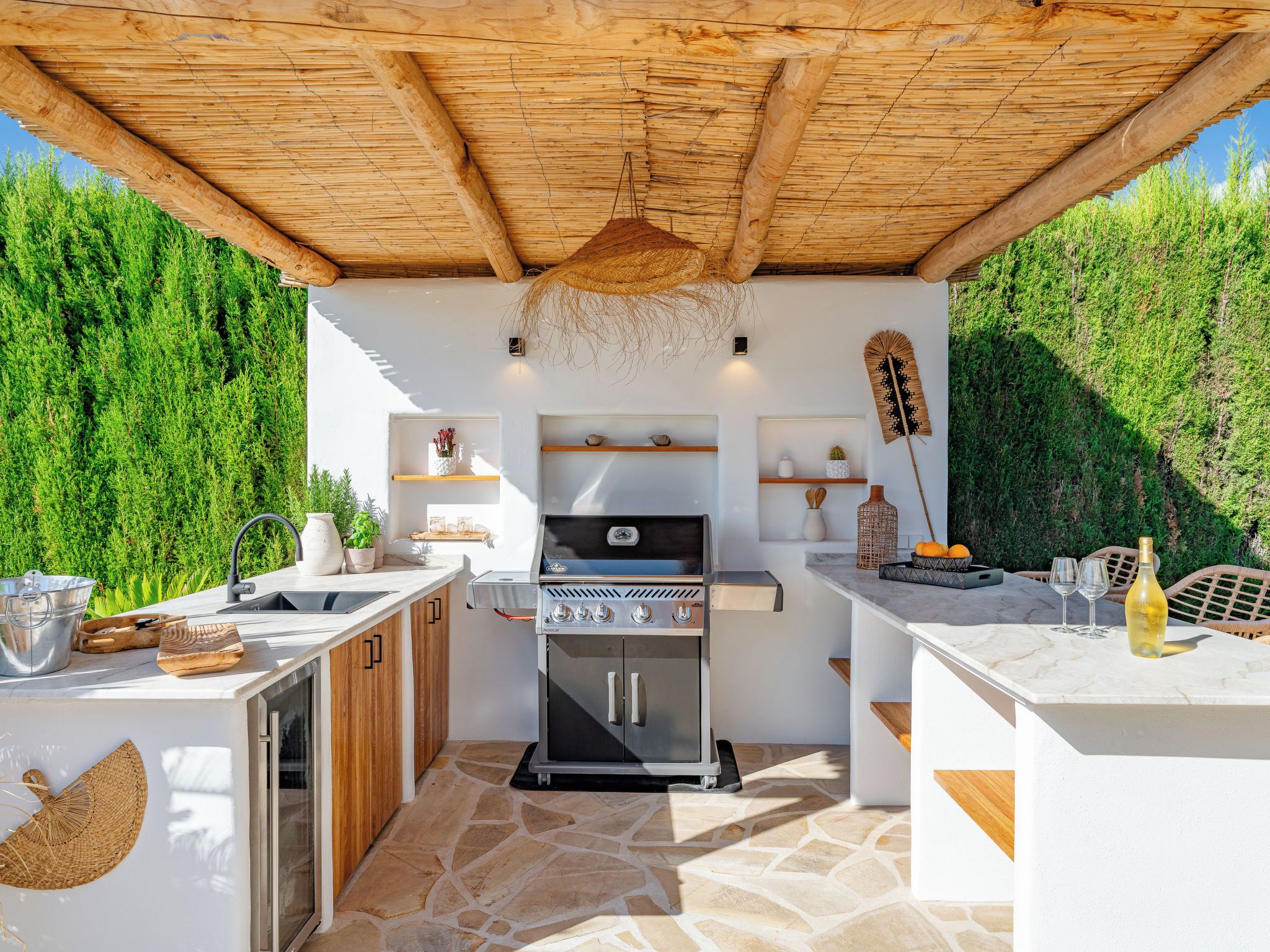Eine Außenküche mit integriertem Grill, Steinboden und einer Pergola darüber.