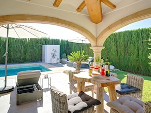 Zona junto a la piscina con mobiliario de mimbre, una mesa con botellas y vistas a una piscina y follaje.