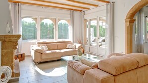 A spacious living room with a beige sofa, a glass coffee table, and a fireplace. Large windows offer views of greenery outside. - Villa SanMar by Interhome (Jávea)