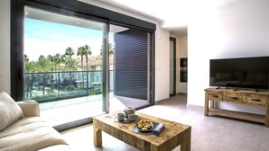 Una sala de estar moderna con un gran ventanal corredera que da a un balcón, una mesa de centro de madera con objetos decorativos, un televisor de pantalla plana sobre un mueble de madera y un sofá color beige.