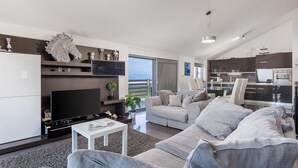 Een moderne woonkamer met een grote flatscreen-tv, een grijze bank, een kleine salontafel en uitzicht op het groen door een schuifdeur van glas.