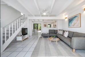 A modern living room with a grey sectional sofa, a flat-screen TV on a white stand, a staircase with white railings, and a ceiling fan. - 'Villa One' Walk to Noosa Beach & Hastings Street (Noosa)