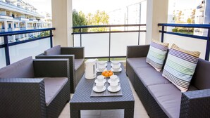 En balkon med fletmøbler, et bord dækket med hvide kopper og underkopper, samt udsigt til et krydstogtskib og bygninger.