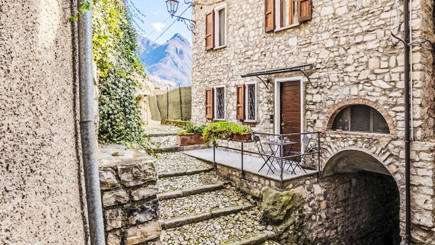 Un bâtiment en pierre avec un petit espace extérieur aménagé en coin salon, un escalier menant à une porte, et une montagne en arrière-plan.