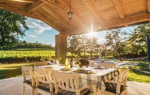Un espace de restauration en plein air avec une table dressée pour quatre personnes, entourée de chaises, sous une pergola en bois agrémentée d’une lanterne suspendue.