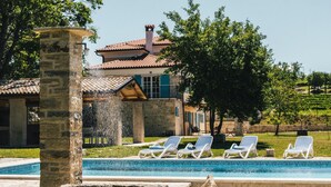A pool area with lounge chairs, a stone pillar, and a house with a tiled roof in the background. - Beautiful home in Buje (Buje)