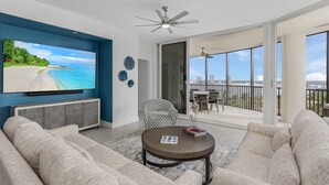 Een ruime woonkamer met een grote flatscreen-tv, een plafondventilator en een balkon met uitzicht op de stad.
