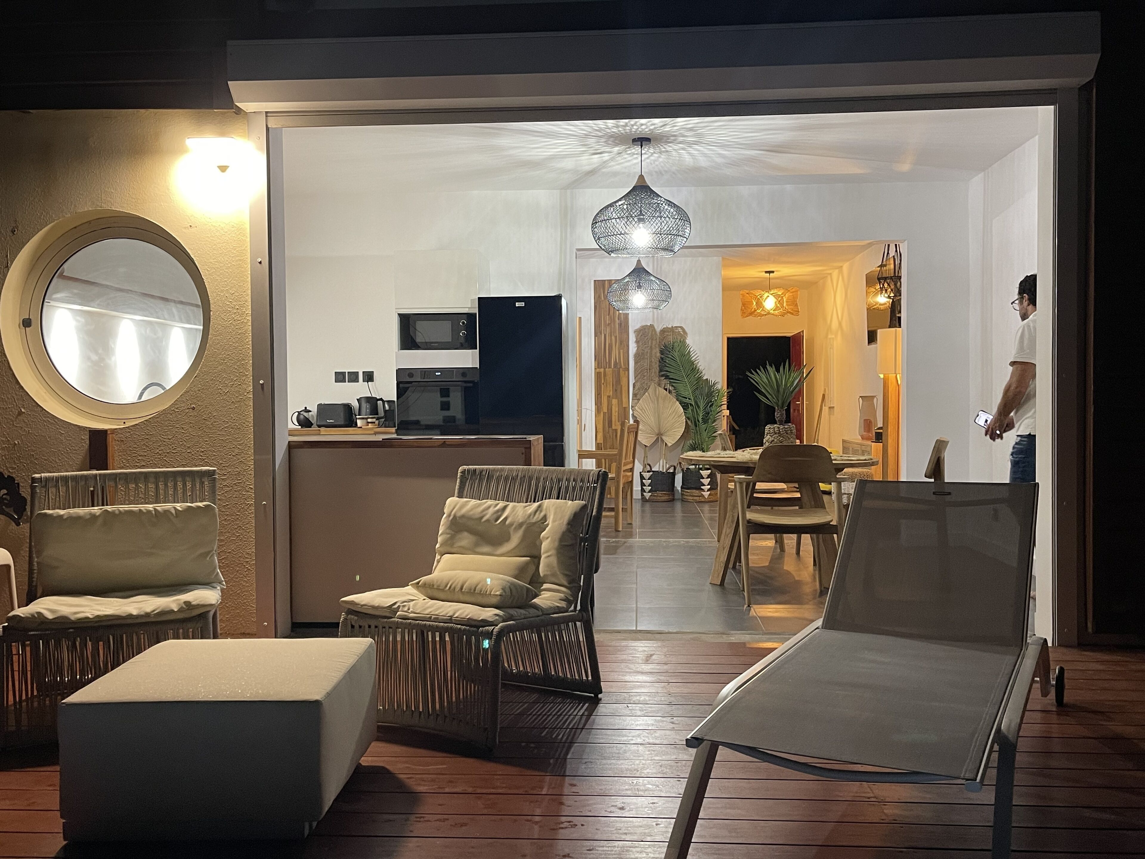 Un salon moderne avec des meubles en osier, un miroir rond et un coin repas comprenant une table et des chaises.
