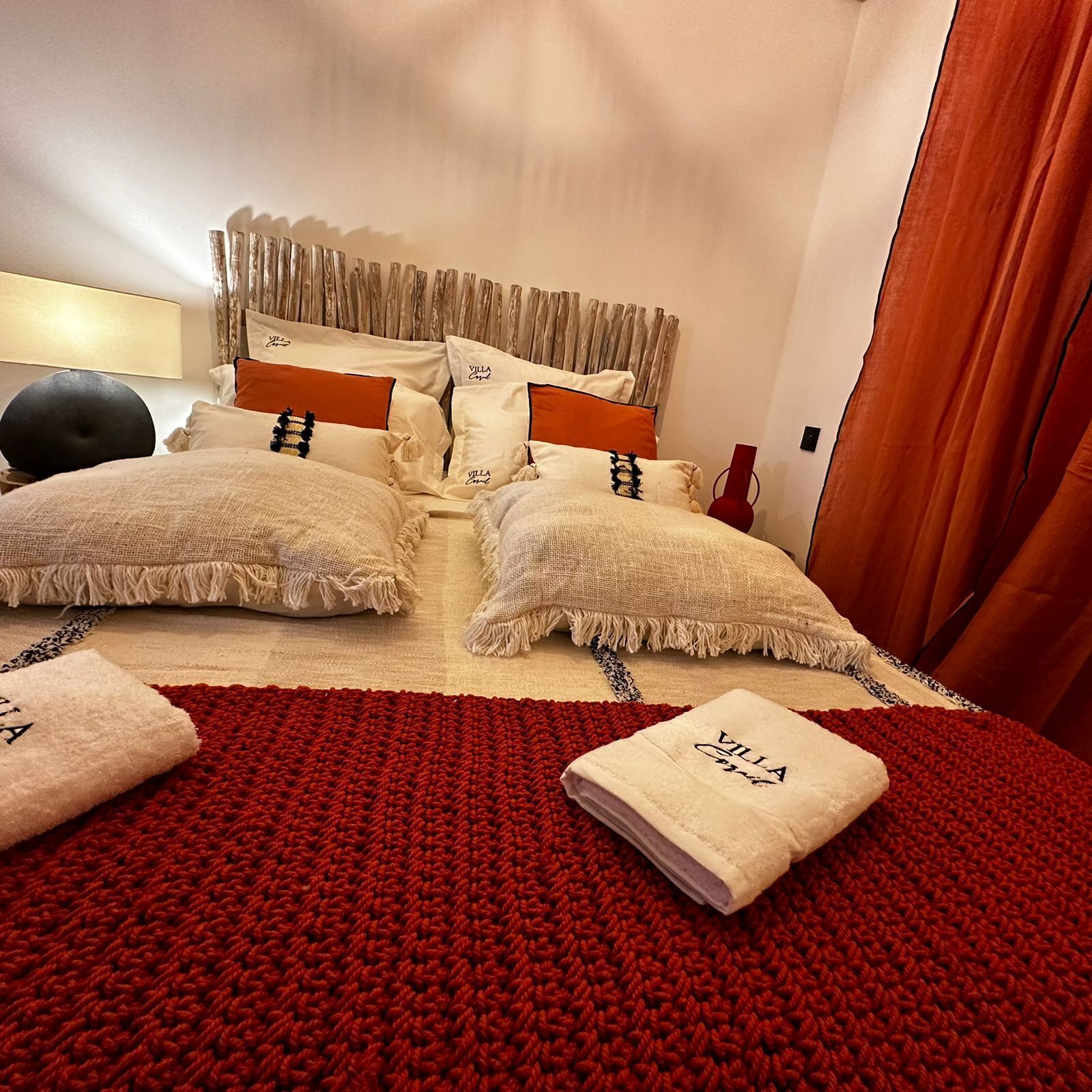A neatly made bed with a red blanket, two pillows, and a folded towel on a red knitted rug.