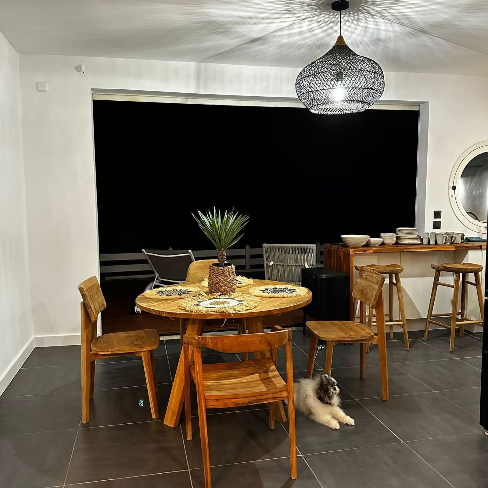 A dining area with a round wooden table, chairs, a hanging light fixture, and a dog lying on the floor.