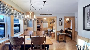 L’intérieur chaleureux d’un chalet comprend un coin repas avec des meubles en bois et un lustre. Un téléviseur est fixé au mur, et divers éléments de décoration sont présents, notamment un poisson peint sur le mur et une enseigne sur laquelle on peut lire « MAISON AU BORD DU LAC ».