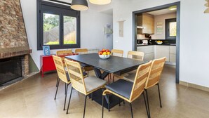 Ein Essbereich mit einem schwarzen Tisch, Korbstühlen und einer Obstschale. Ein Kamin aus Stein, ein roter Schrank und eine Küche sind durch einen offenen Durchgang sichtbar.
