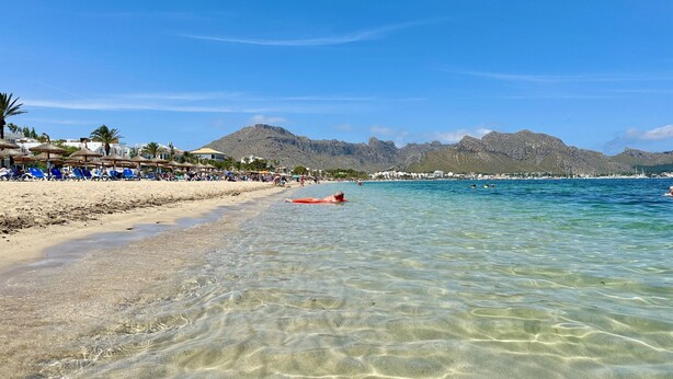 A beach with clear shallow waters, a person in the water, and mountains in the background.