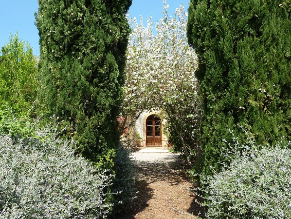 Uma trilha leva a um edifício com entrada em arco, cercado por árvores perenes e arbustos floridos.