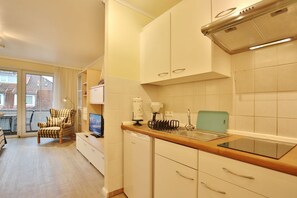 A compact kitchen with a sink, a microwave, and a small countertop. There is a chair and a TV in the adjacent living area.