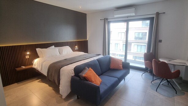 A modern hotel room with a large bed, a sofa, a small table, and a view of the city through a large window. - Chic Studio in The Signature, Accra (Accra)