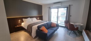 A modern hotel room with a large bed, a sofa, a small table, and a view of the city through a large window. - Chic Studio in The Signature, Accra (Accra)