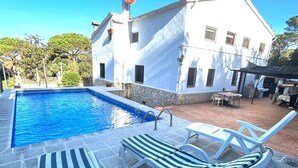A swimming pool with two lounge chairs and a table, surrounded by a stone patio and a white building with windows.
