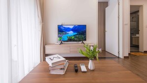 Moderni olohuone, jossa on jalustalla seisova taulutelevisio, sohvapöytä, jossa on kirjoja ja kaukosäätimiä, ruukussa oleva huonekasvi ja puulattia.