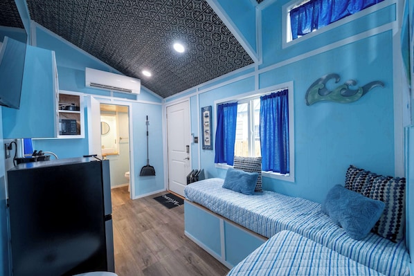 A compact room with a kitchenette, a bed with patterned bedding, a ceiling with a decorative design, and a window with blue curtains.
