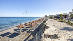 A beach with rows of sun umbrellas and lounge chairs, a sandy shore, and a building with a modern design in the background.